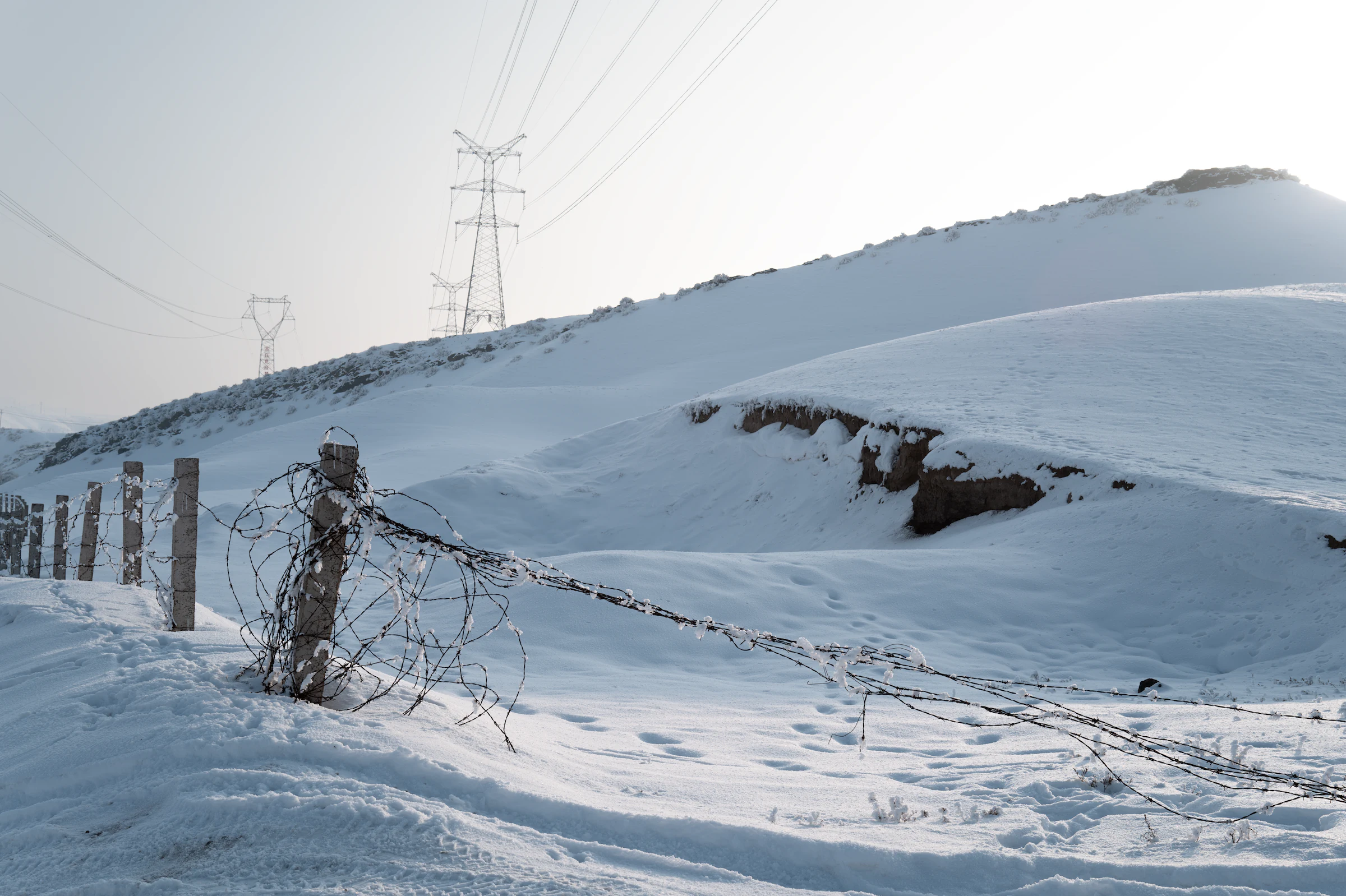 新疆乌鲁木齐的厚雪覆盖坡地与围栏，输电线沿山坡延伸